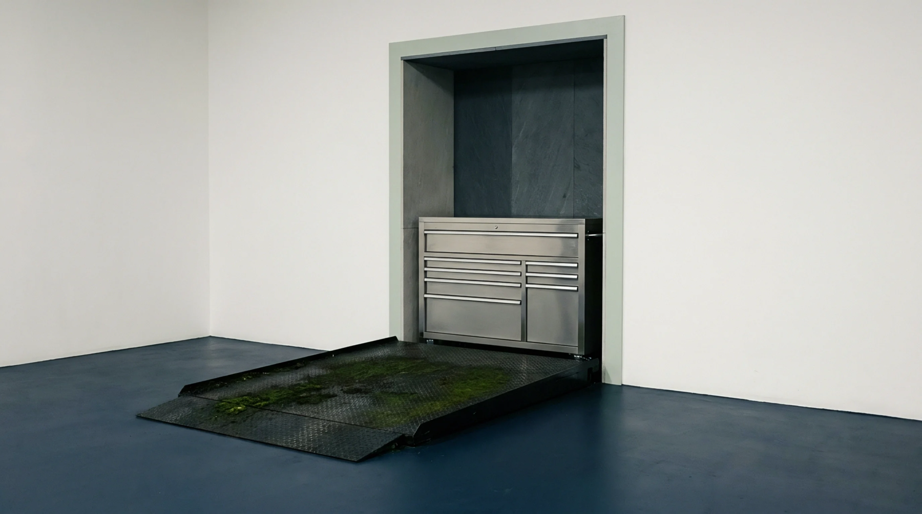 Stainless steel storage cabinet sits within a recessed concrete wall beside a weathered steel loading ramp with organic growth.