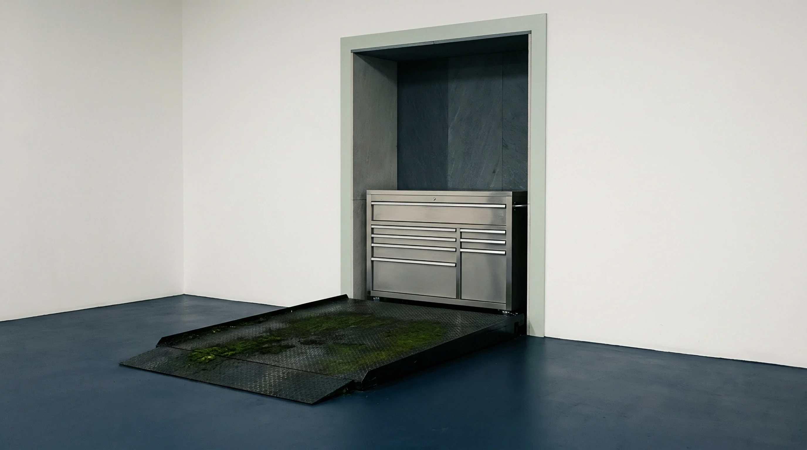 Stainless steel storage cabinet sits within a recessed concrete wall beside a weathered steel loading ramp with organic growth.