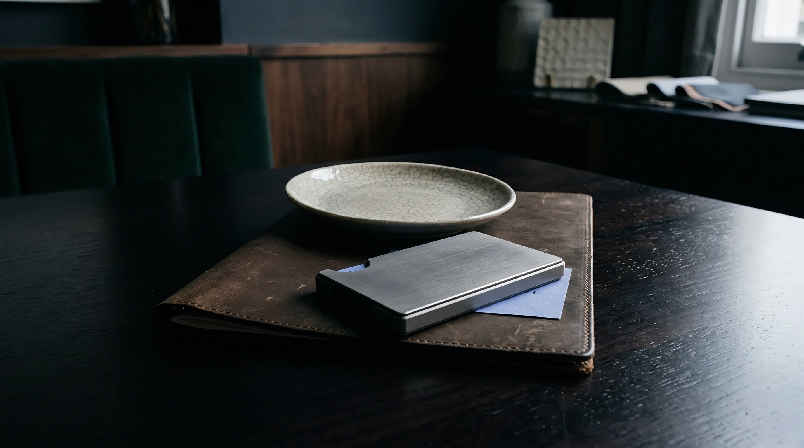 A leather procurement folder and metal cardholder rest on a dark wood table beside a ceramic product sample plate.